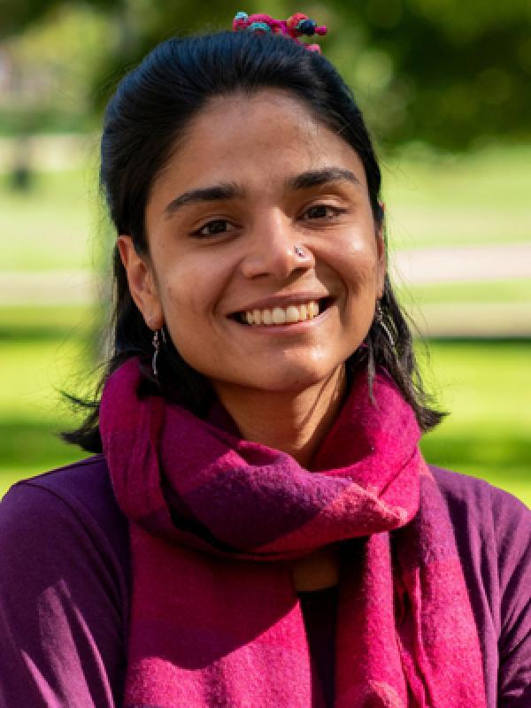  This photo features Paromita Bathija smiling outdoors. She is wearing a purple top with a matching pink scarf, and her hair is tied back with a colorful hair accessory. The background is a lush, green park area, creating a bright and inviting atmosphere.