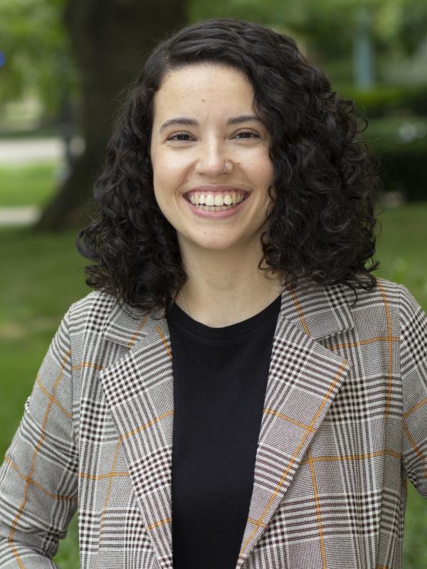 This photo features Taís Souza Carareto smiling outdoors. She has curly dark hair and is wearing a black shirt under a plaid blazer. The background is a lush, green park area, and she conveys a bright and cheerful demeanor.