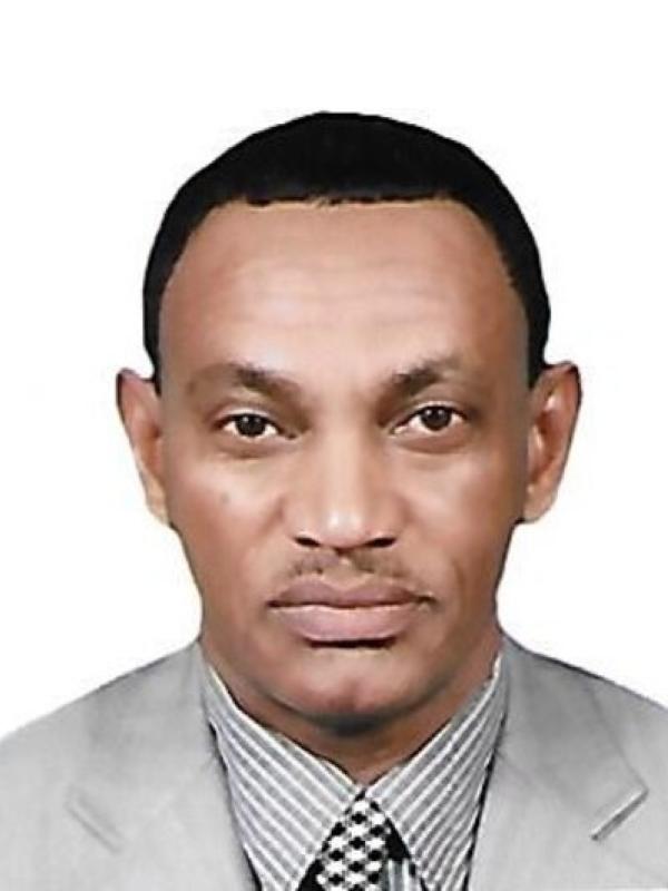 This photo features Ahmad Sikainga in front of a plain white background. He is wearing a light gray suit jacket over a patterned shirt and a checkered tie. His expression is calm and professional, and he has neatly trimmed hair.