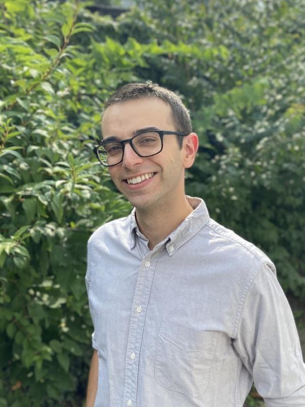 This photo features Jared Rabinowitz smiling outdoors. He is wearing glasses and a light gray button-down shirt. The background is filled with lush green foliage, and the overall atmosphere is bright and cheerful.