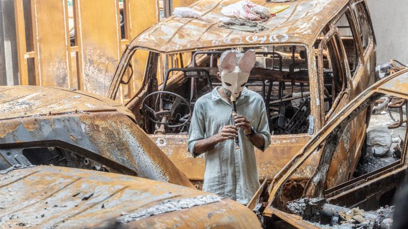 Individual wearing rabbit mask playing recorder surrounded by rusted cars