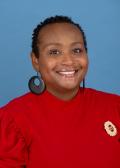 A smiling Le'Trice Donaldson, wearing a red top with a brooch and large black earrings, poses against a blue background.