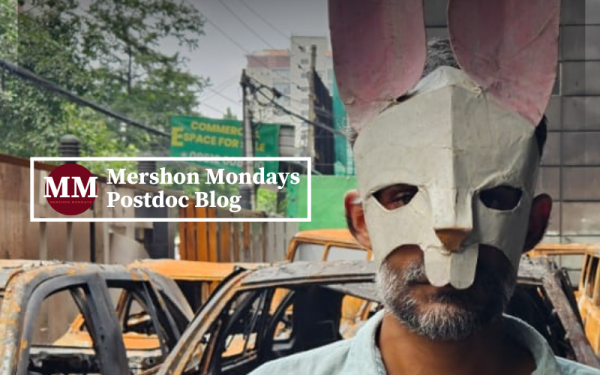 Mershon Monday logo over image of man with rabbit mask standing in front of rusted vehicles