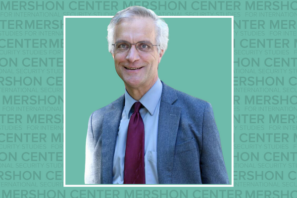 An image of a man with gray hair wearing glasses and a suit, standing before a green background with the Mershon Center logo pattern