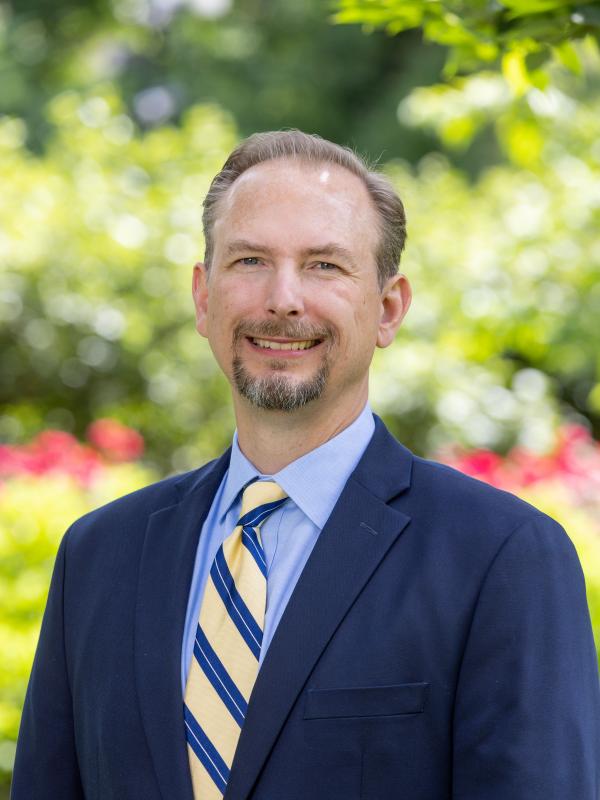 Dakota Rudesill in a navy suit, light blue dress shirt, and yellow and blue striped tie stands outside.