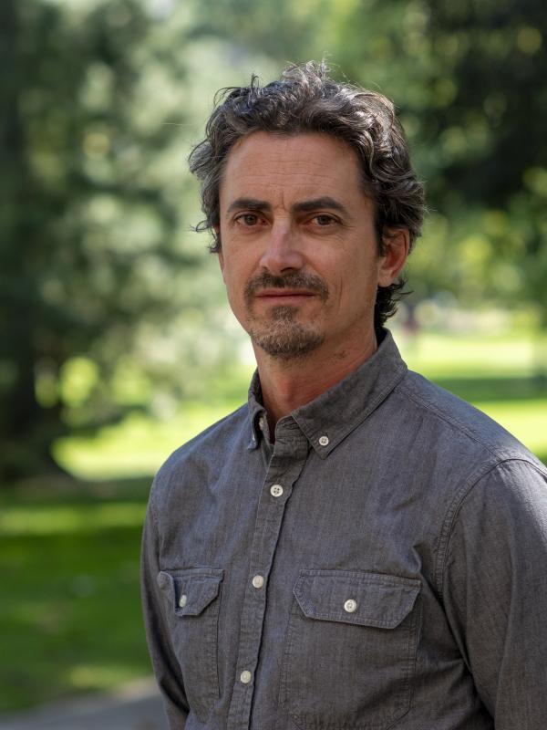  This photo features Max Woodworth standing outdoors and smiling slightly. He is wearing a dark gray button-down shirt and has curly gray hair. The background is a bright, green park area, providing a relaxed and natural atmosphere.