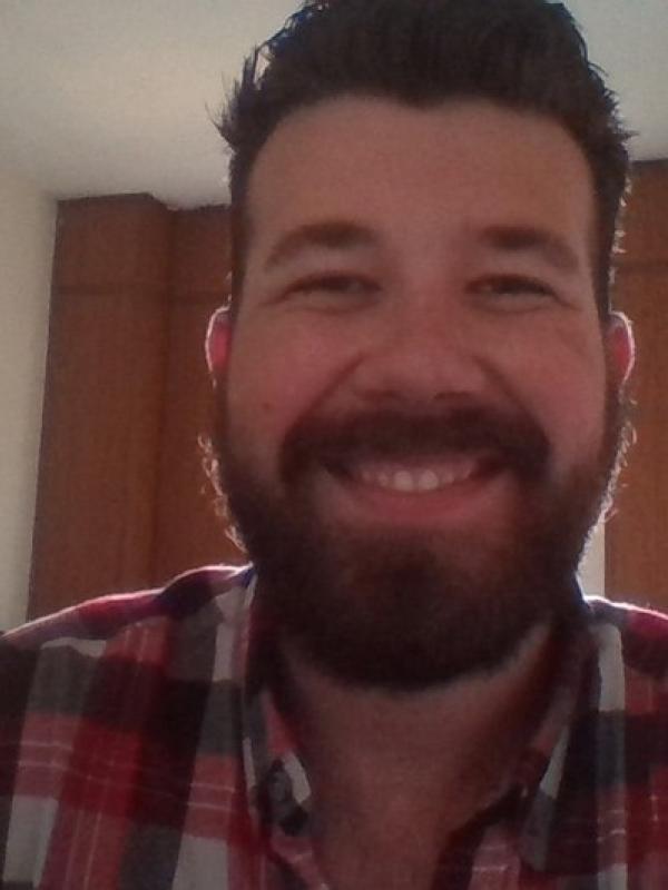 This photo features Ian Gammon smiling indoors. He has a full beard and is wearing a red plaid shirt. The background shows wooden cabinets and soft lighting, giving the image a warm and relaxed atmosphere.