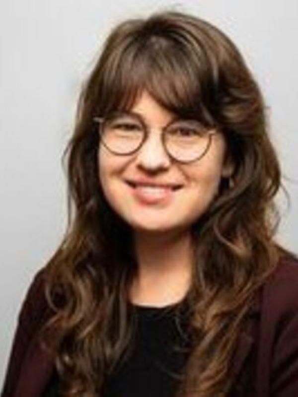 This photo features Erin Moore smiling against a plain gray background. She is wearing round glasses, a black top, and a maroon blazer. Her hair is long and wavy.